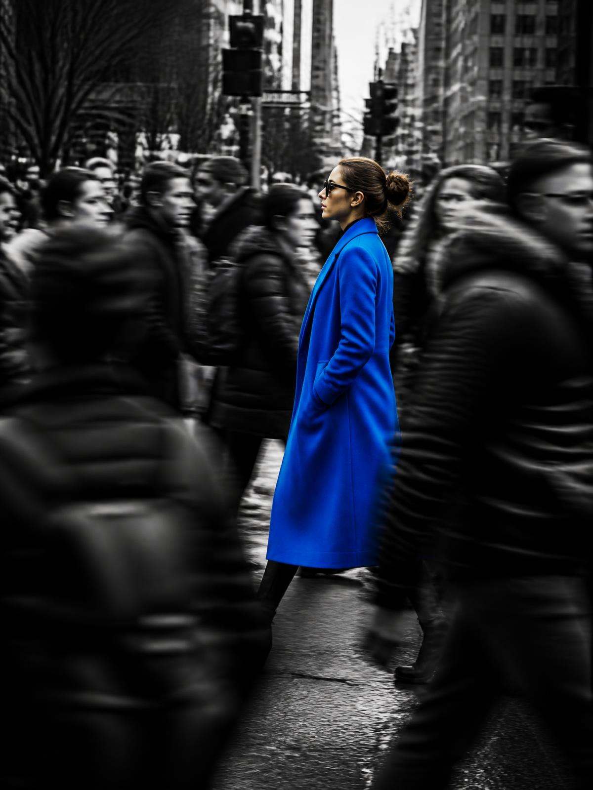 A person in a bright blue coat standing still in a rushing black-and-white crowd